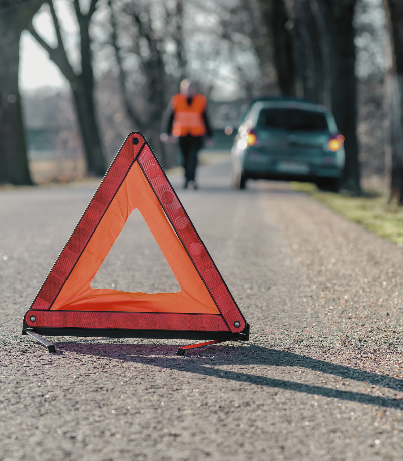 Persona a lo lejos con chaleco reflectante naranja al lado de un coche averiado y en primer plano triángulo de señalización.
